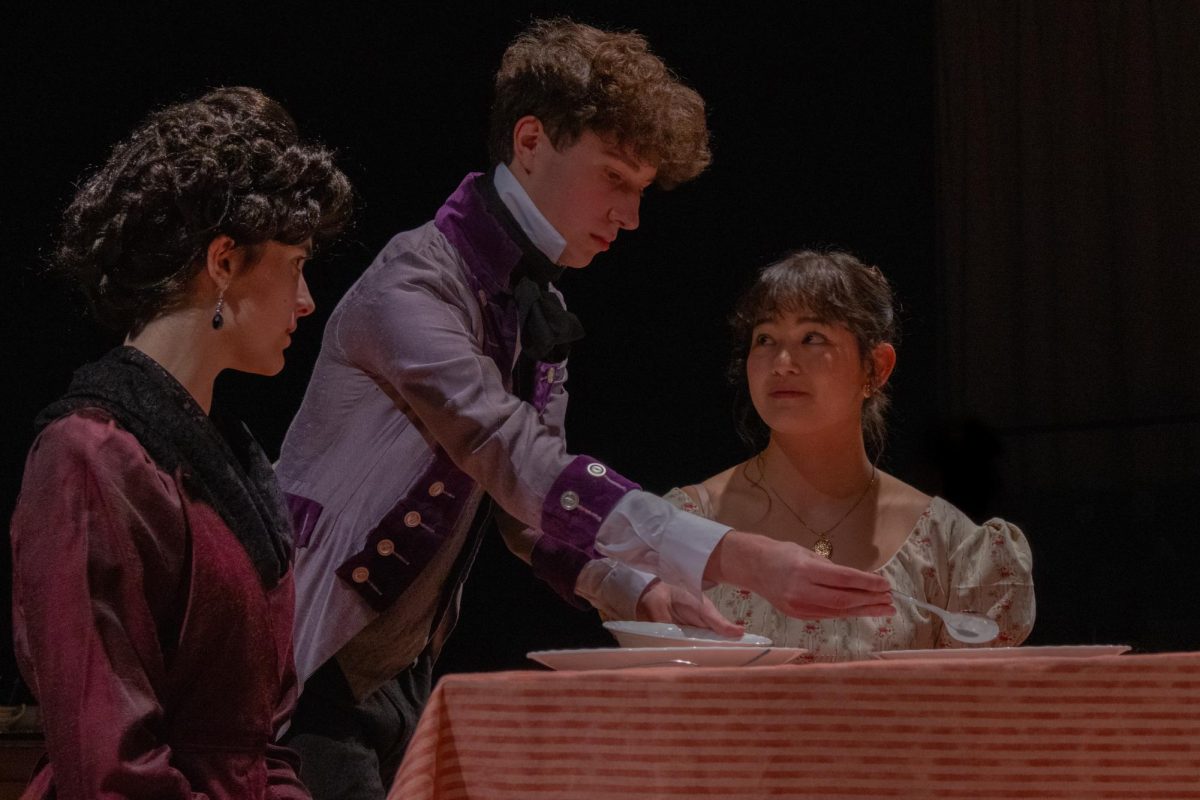 Amidst dining room set pieces, Adam Trenk '26 - as Thomas - pantomimes serving food to fellow actors Saffron Kollodge '26 and Sabrina Kim '26. Trenk was one of the actors to play a "Gossip," members of the ensemble who, in addition to performing roles like Thomas, also served as narration for the show. They bookended scenes and spoke to audience members, representing the gossip in society surrounding the play's events.