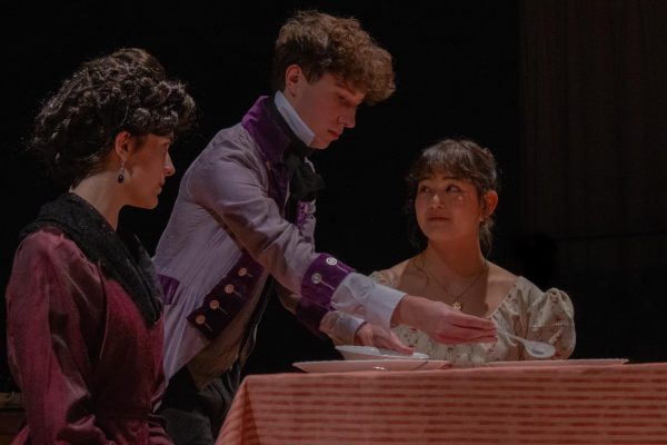 Amidst dining room set pieces, Adam Trenk '26 - as Thomas - pantomimes serving food to fellow actors Saffron Kollodge '26 and Sabrina Kim '25. Trenk was one of the actors to play a "Gossip," members of the ensemble who, in addition to performing roles like Thomas, also served as narration for the show. They bookended scenes and spoke to audience members, representing the gossip in society surrounding the play's events.