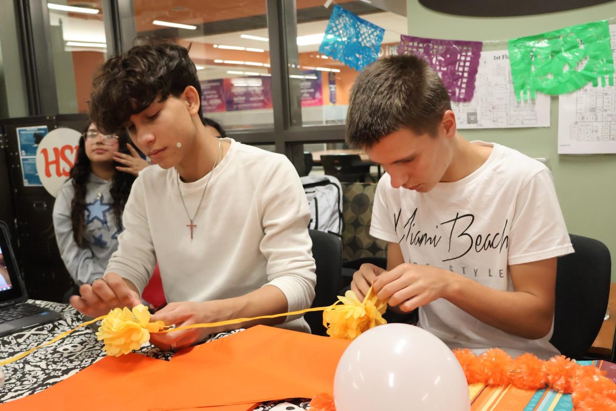 Concentrated and determined, Miguel Torres '29 and Diego Rey '28 cut out and tie paper marigolds to create a garland. Along with surrounding students, they followed directions from a crafting video displayed on the screen to create the craft. "I don't celebrate Día de Los Muertos myself, but I'm glad to be a part of this [event] and celebrate it here," Rey said.