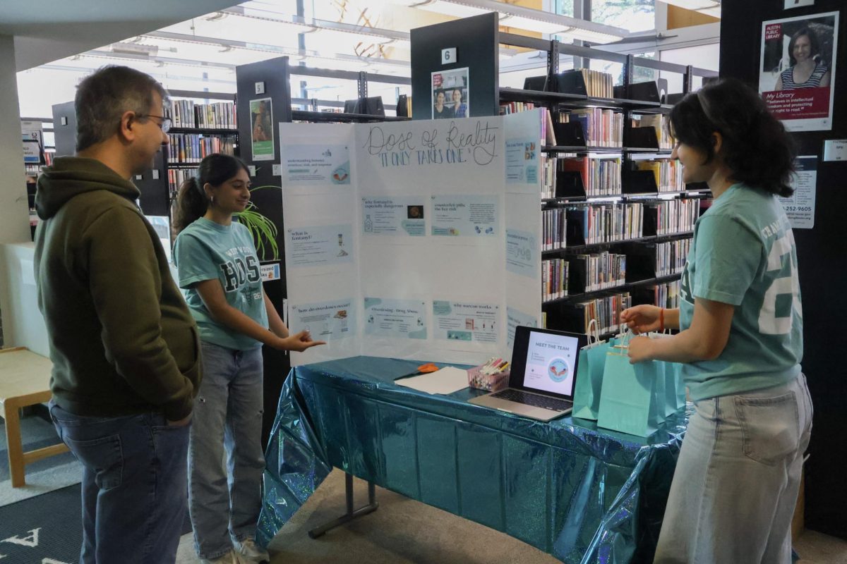 Pointing towards their booth, sophomores Aanya Pathak and Saloni Chitari talk to a library member about show their research and care kits. The students created care kits to give to library members who created a card. 
