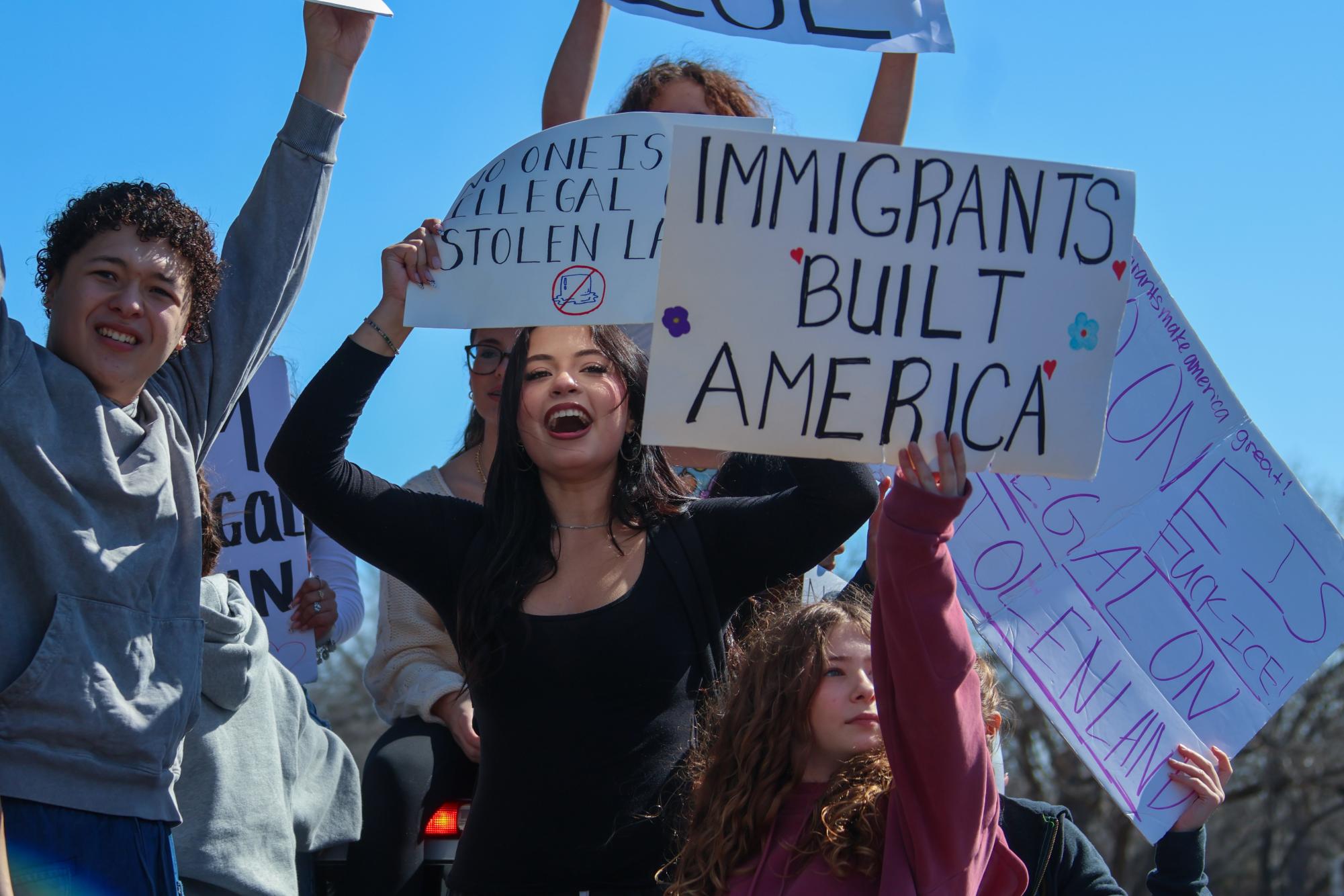 Standing in the bed of a pickup truck, Jillian Huston '26 holds a sign reading "No One is Illegal on Stolen Land" while protesting alongside students on the sidewalk. This phrase argues that since the U.S. was unlawfully seized from Indigenous tribes through broken treaties and forced relocation, the country lacks the moral authority to deem migrants "illegal" on the territory.